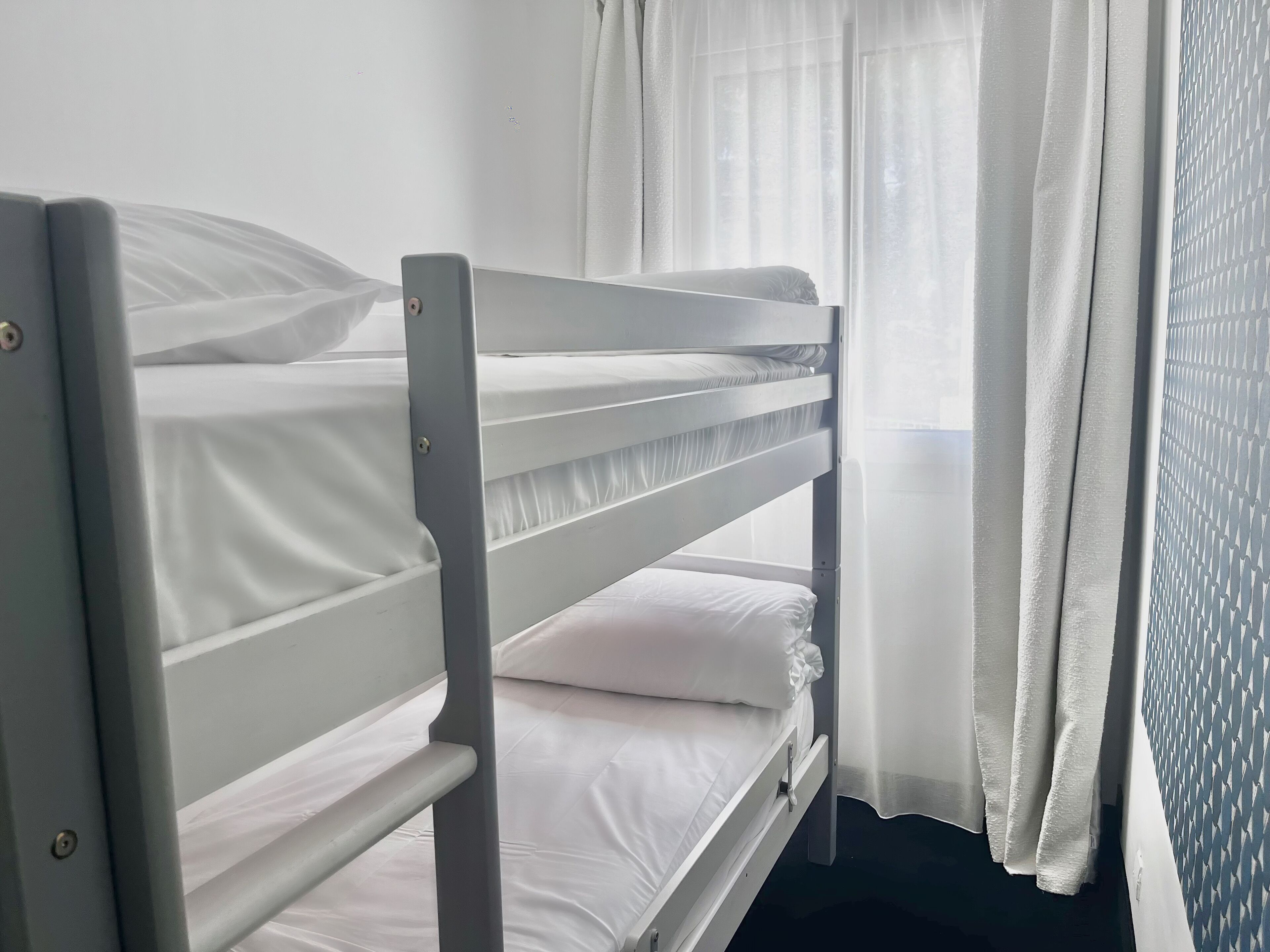 Una cama litera con ropa de cama blanca y una ventana con cortinas traslúcidas.