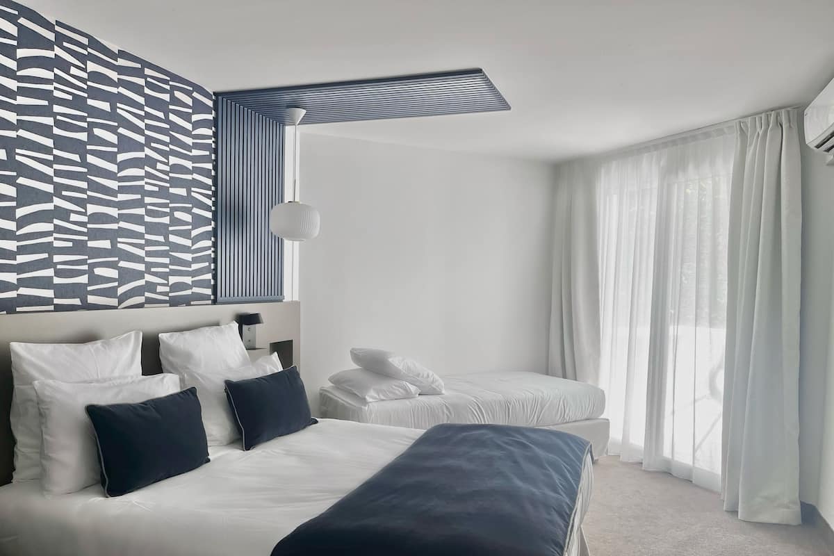 A modern hotel room with a large bed, a smaller bed, and a bench. There is a patterned wall and a window with sheer curtains.