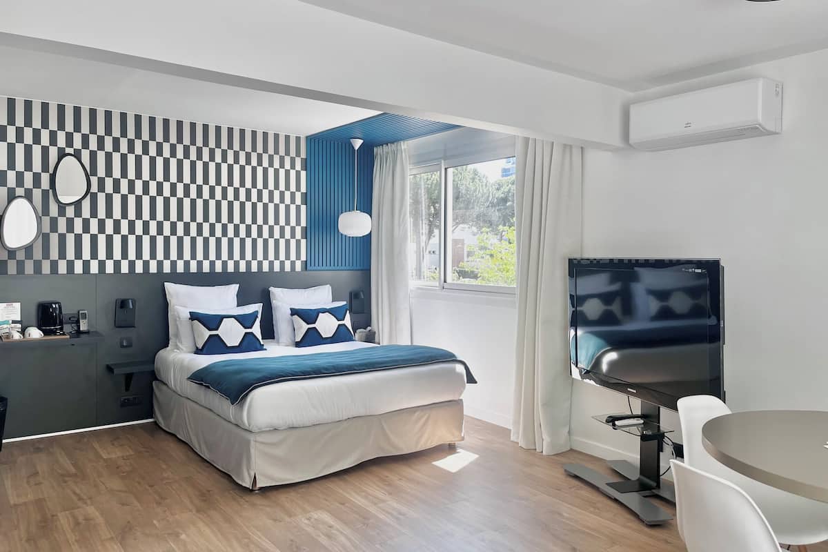 A modern hotel room with a large bed, a desk with a flat-screen TV, a desk lamp, and a window with curtains.