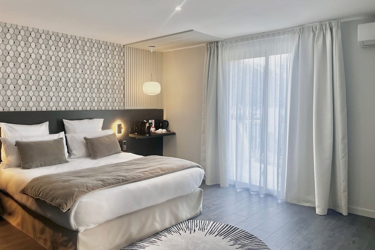 A modern hotel room with a large bed, a nightstand with a lamp, and a view of the outdoors through sheer curtains.