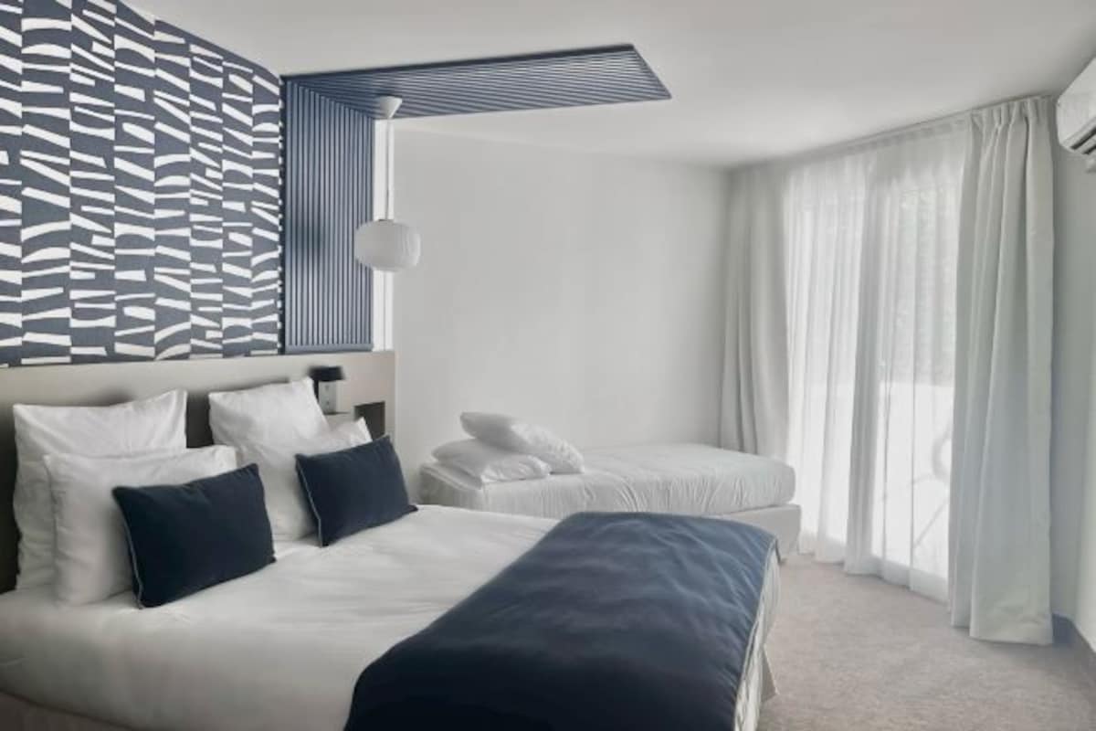 A modern hotel room with a large bed, a smaller bed, a desk, and a patterned wall.