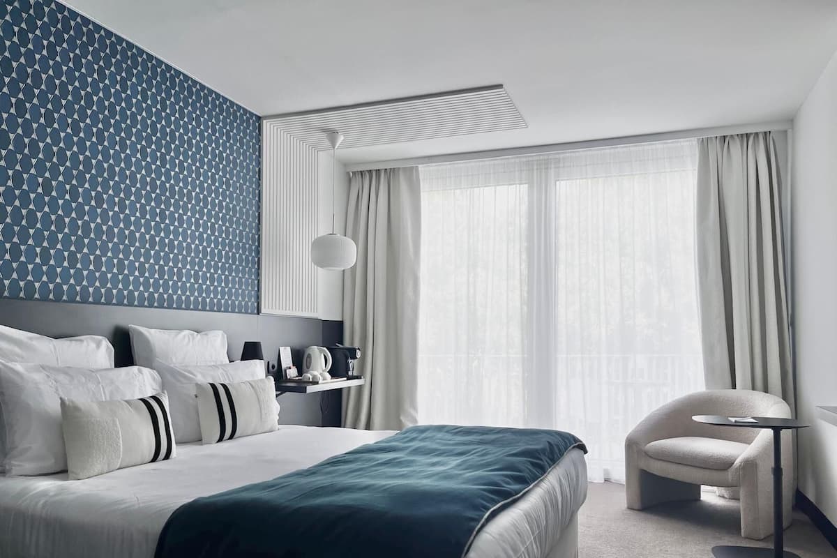 A modern hotel room with a large bed, a bedside table, a chair, and a window with curtains.