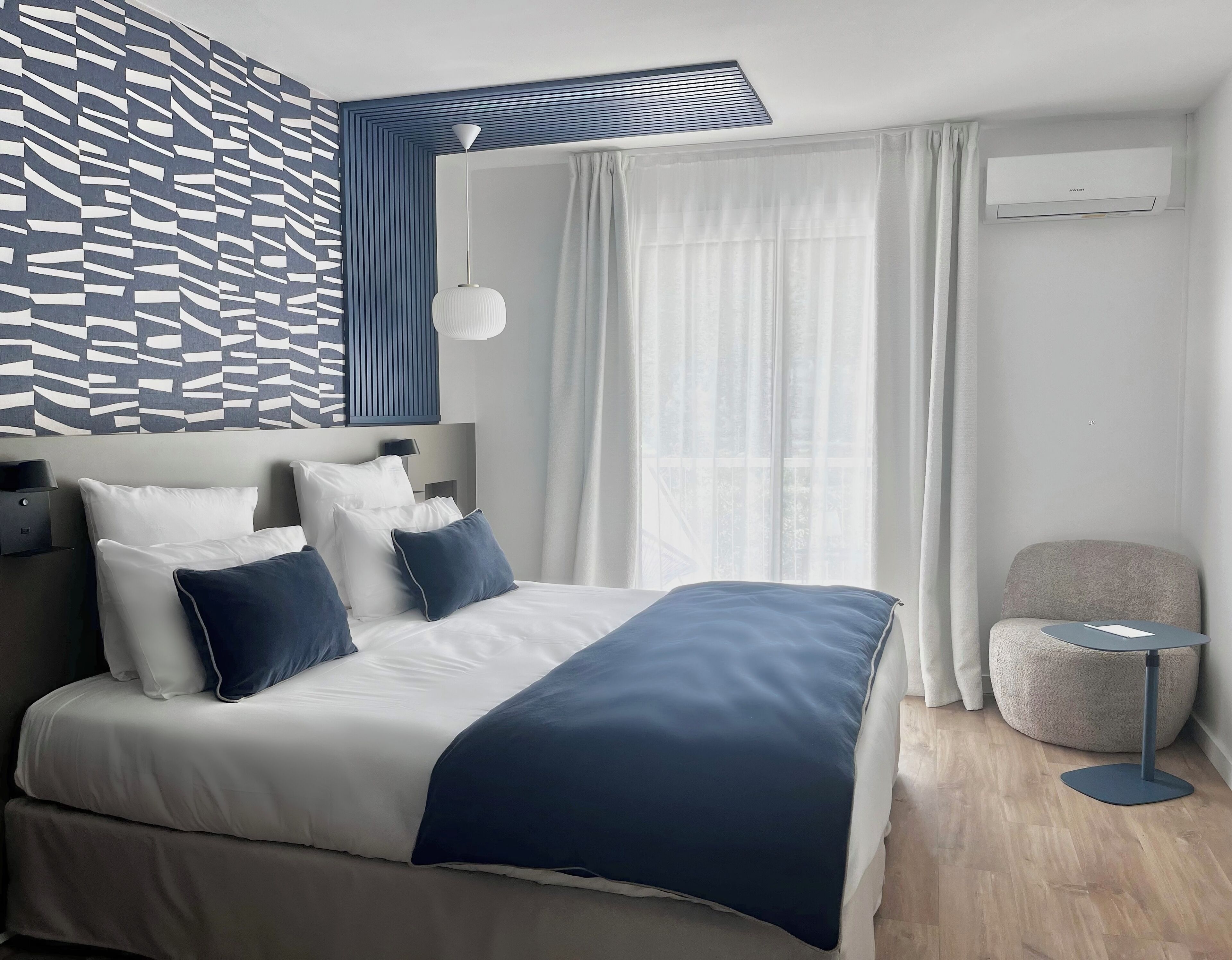 Et moderne hotelværelse med en pænt redt seng, et lille bord, en rund stol og et stort vindue med gardiner.
