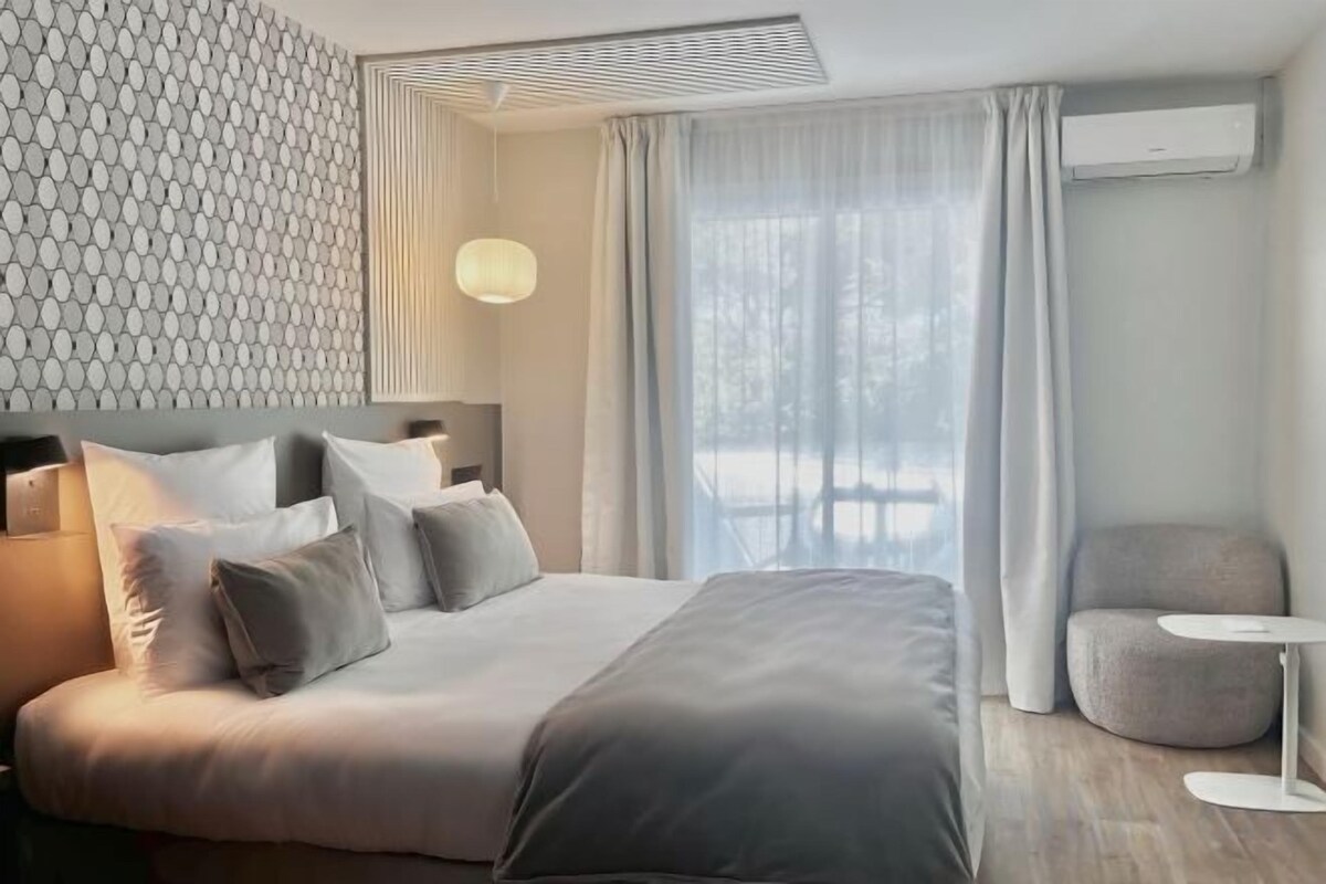 A modern hotel room with a large bed, a bedside table, a chair, and a window with curtains.