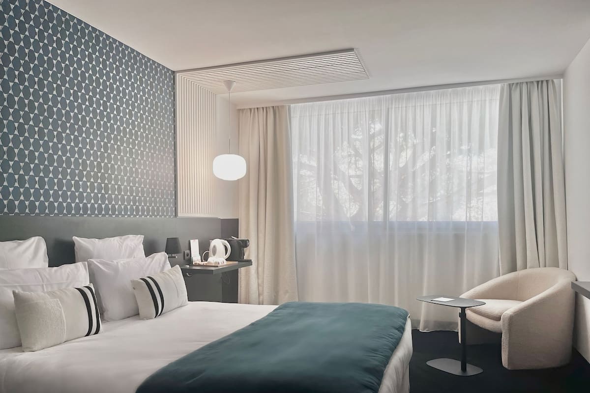 A hotel room with a neatly made bed, a side table with a lamp, a chair, and a large window with sheer curtains.