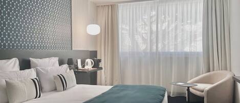 A hotel room with a neatly made bed, a side table with a lamp, a chair, and a large window with sheer curtains.
