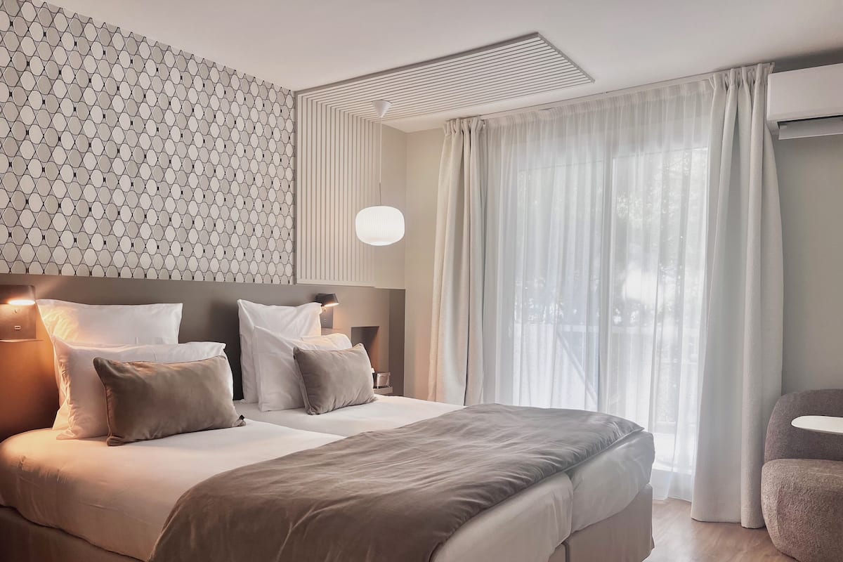 A modern hotel room with a large bed, a patterned wall, a ceiling vent, and sheer curtains.