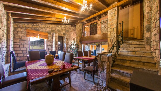 Un comedor rústico con paredes de piedra, vigas de madera y una escalera que conduce a un nivel superior. Las mesas están cubiertas con manteles rojos y sillas.