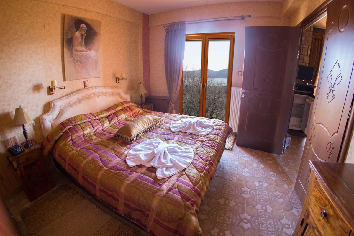 A bedroom with a large bed, a nightstand with a lamp, a painting on the wall, and a view of the outdoors through a door.