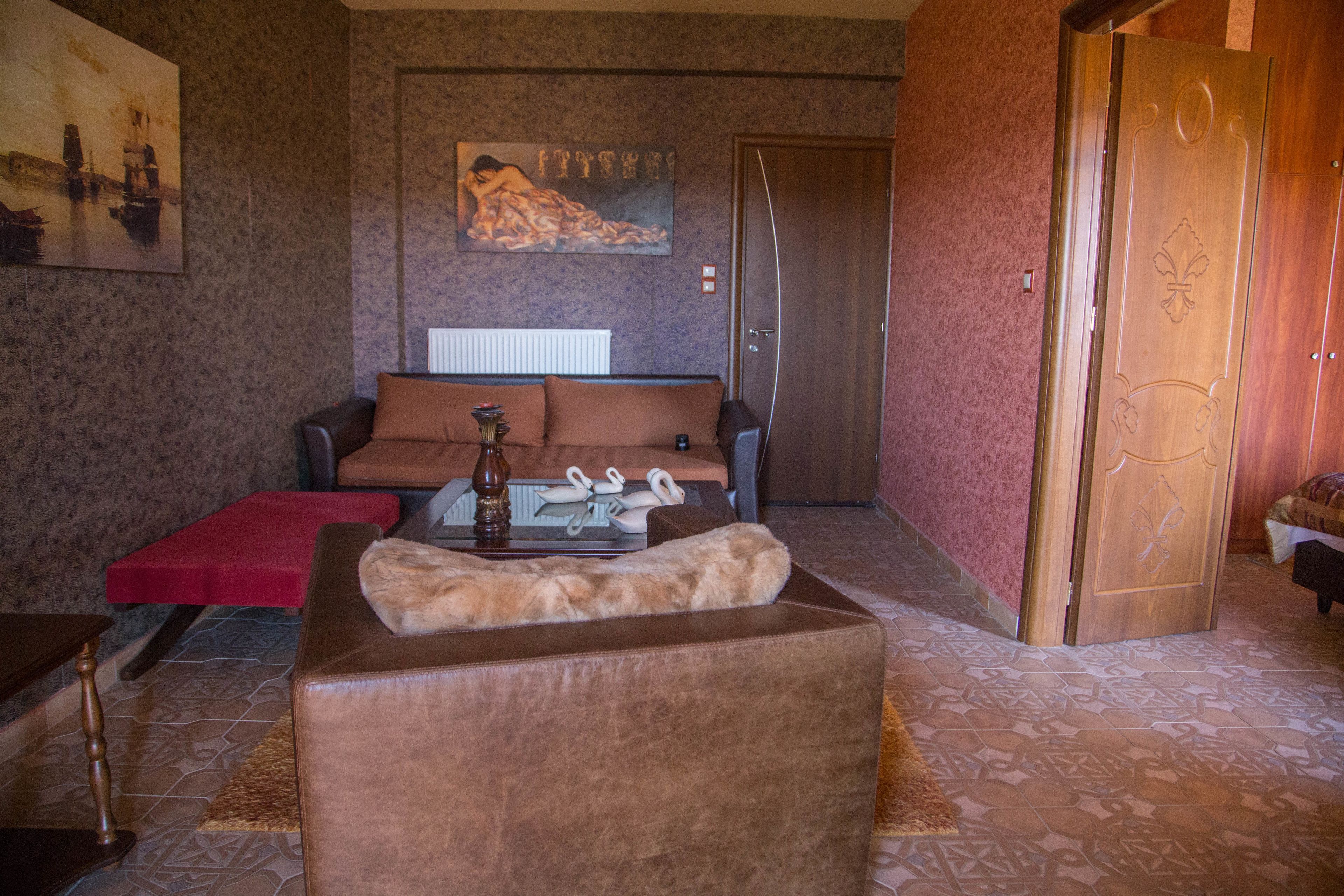 A living room with a brown sofa, a red ottoman, a coffee table with decorative items, a painting on the wall, and a wooden door.