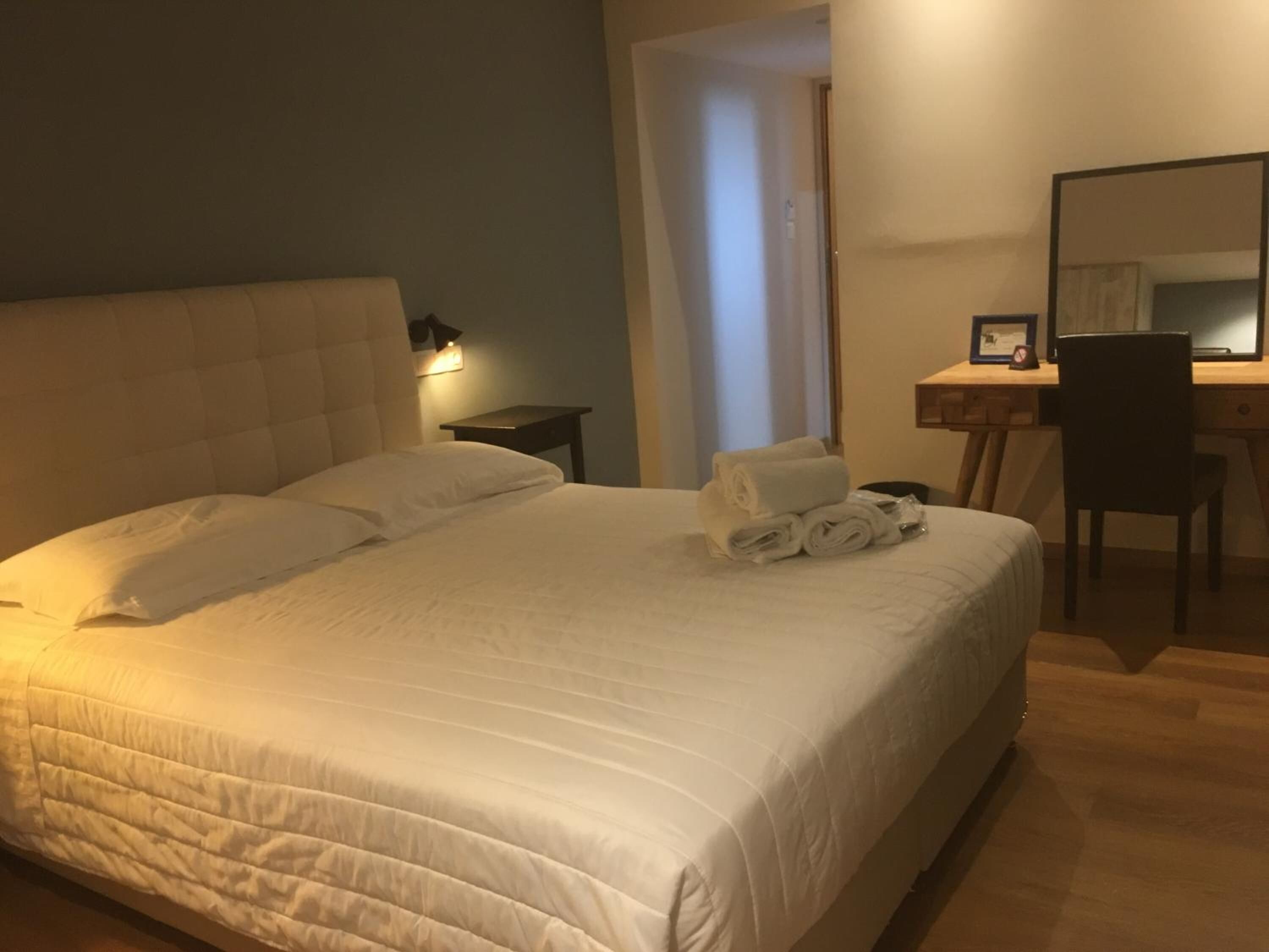 Ein ordentlich bezogenes Bett mit Kissen und gefalteten Handtüchern, ein Nachttisch mit Lampe, ein Schreibtisch mit Stuhl und ein Spiegel.