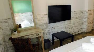 Una habitación con cama, escritorio, silla, mesita, televisor y cubo de basura.