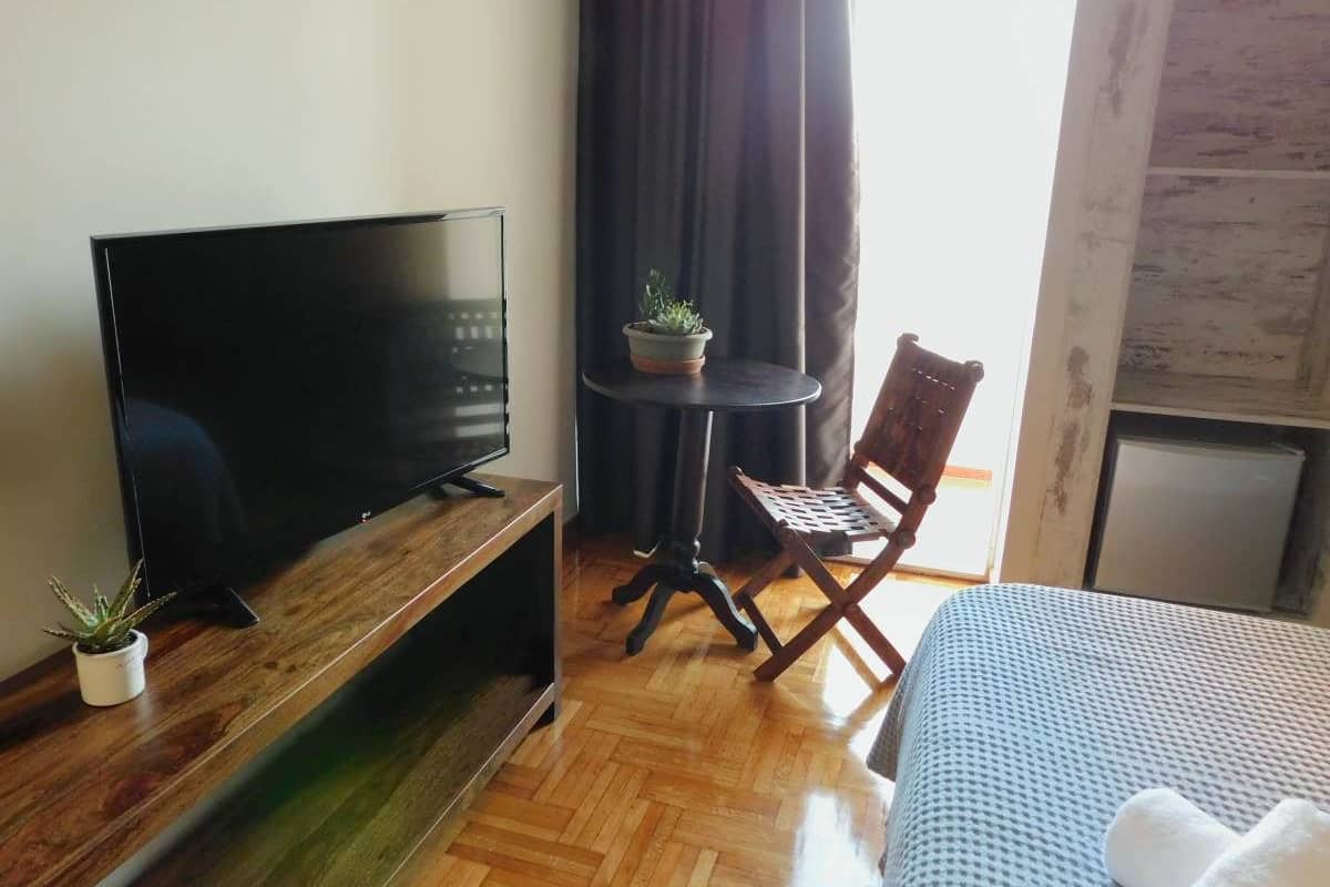 Habitación de hotel con televisor de pantalla plana sobre un soporte de madera, una mesita con planta en maceta, una silla plegable y una cama con cubierta azul.