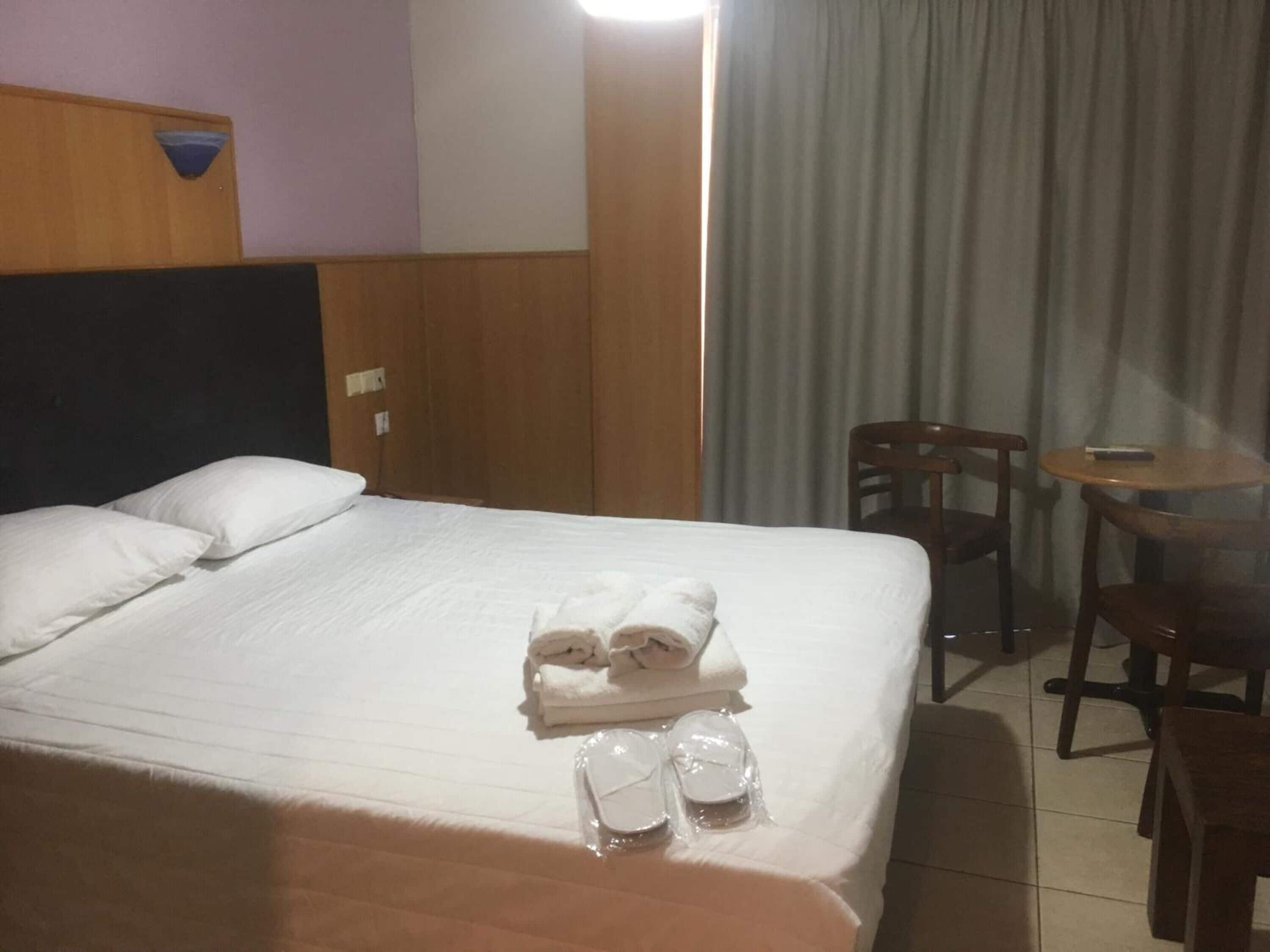 A hotel room with a neatly made bed, a small table with a chair, and a view of the outside through a window with curtains.