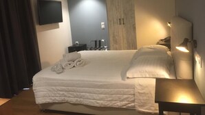 A hotel room with a large bed, a flat-screen TV mounted on the wall, a wooden wardrobe, a bedside table with a lamp, and a small table with a lamp.