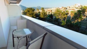 A balcony with a glass table and two chairs, overlooking a town with buildings and trees.