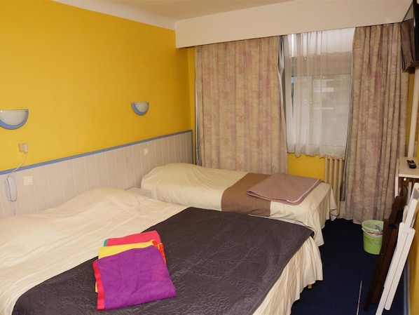 A hotel room with two beds, a small table, a chair, a window with curtains, and a radiator. - Hôtel le Richelieu (Le Boulou)