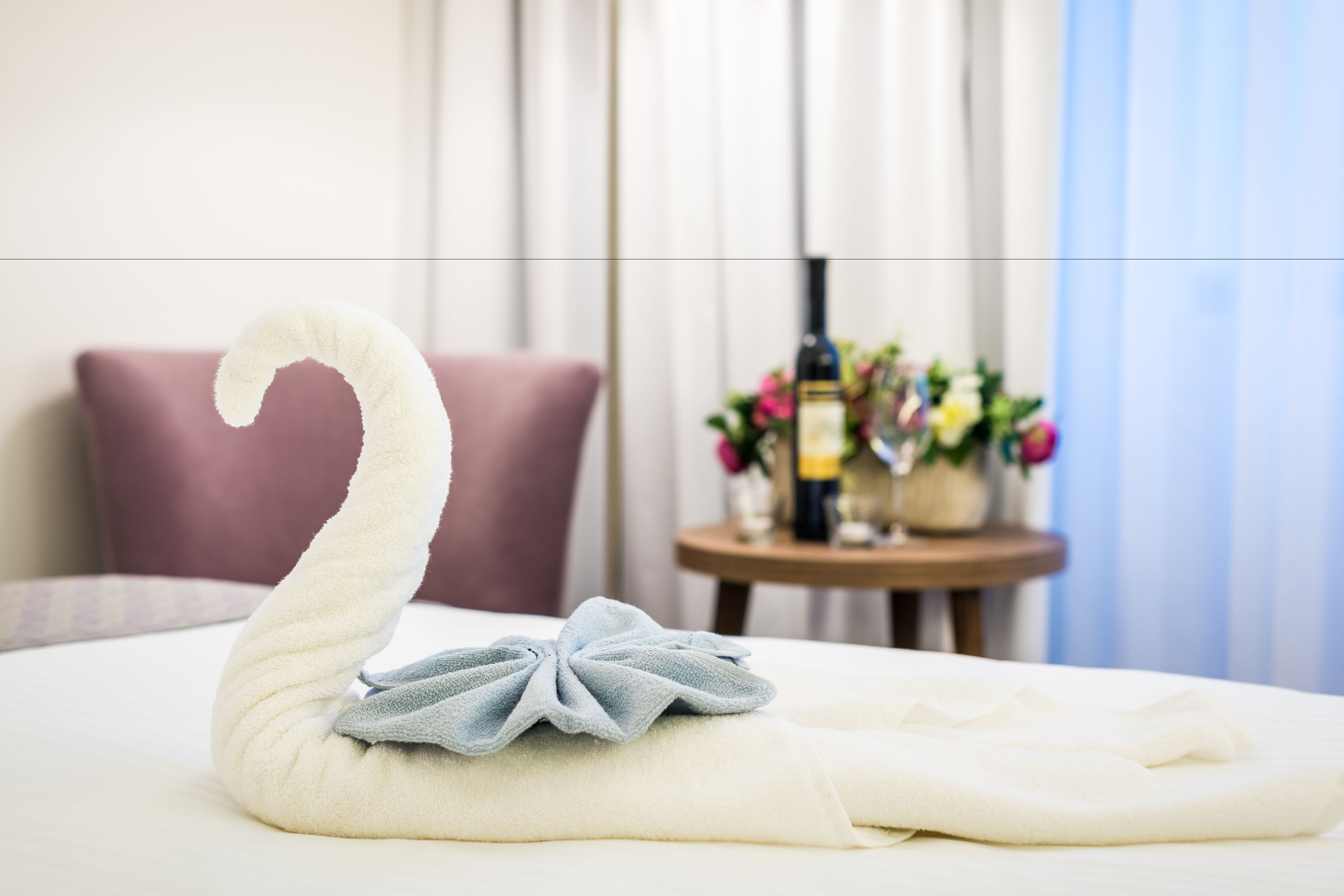 A neatly made swan-shaped towel on a bed with a small round table and a bottle of wine in the background.