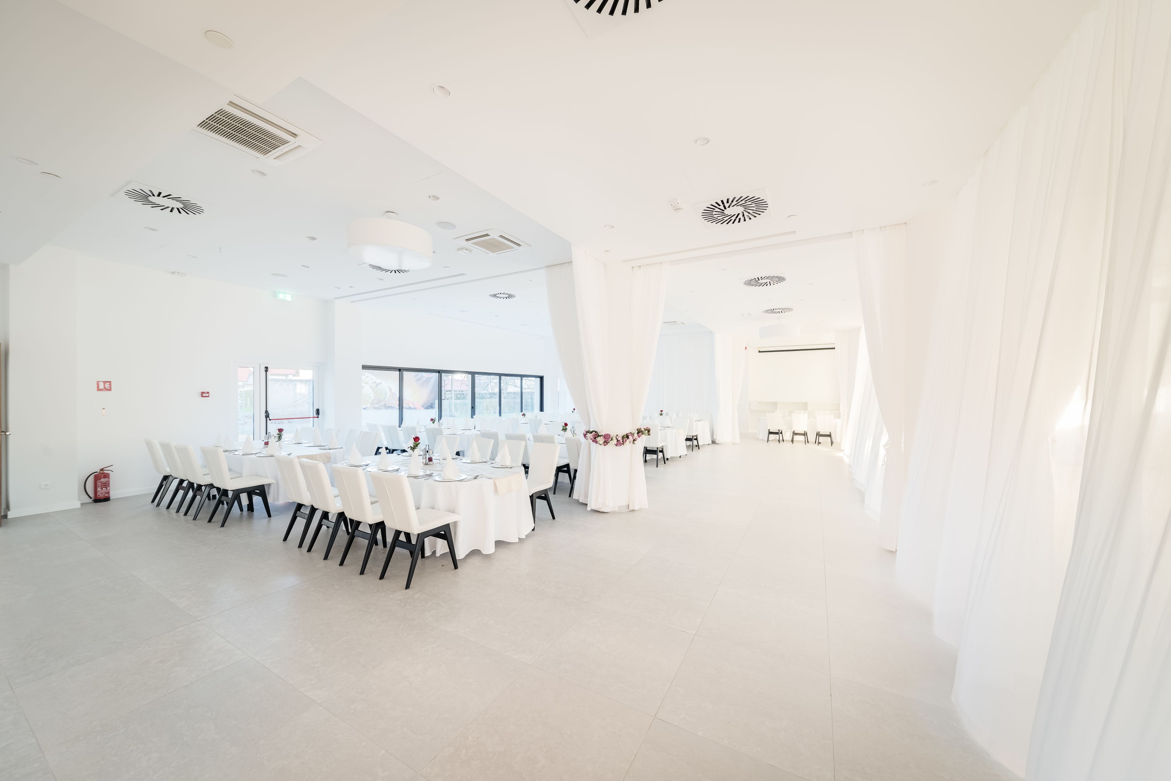 Een ruime, goed verlichte kamer met witte wanden en een hoge plafond, met een lange tafel gedekt voor een formeel evenement.