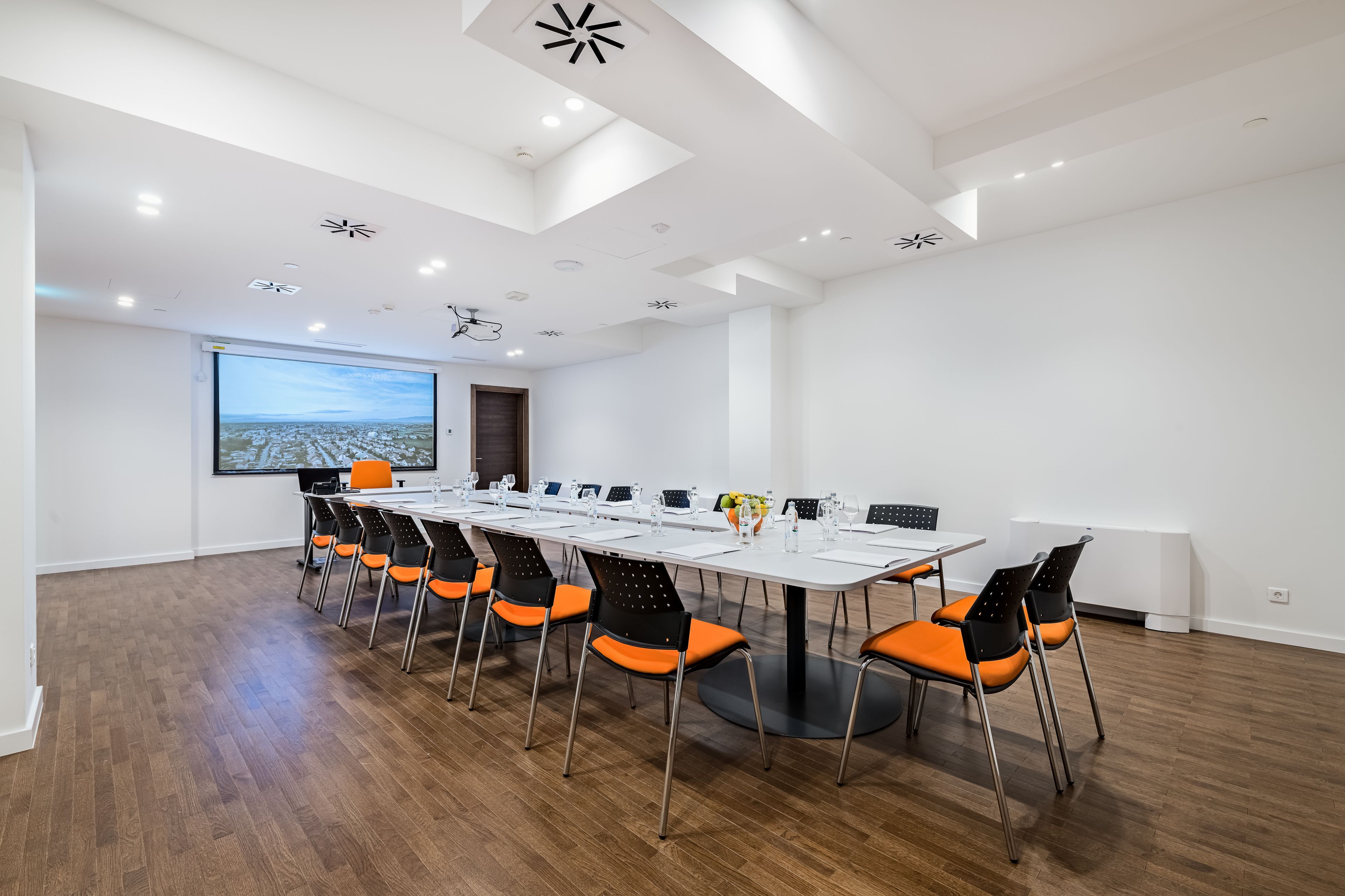 A conference room with a long table, orange chairs, white walls, and a large screen displaying a cityscape.