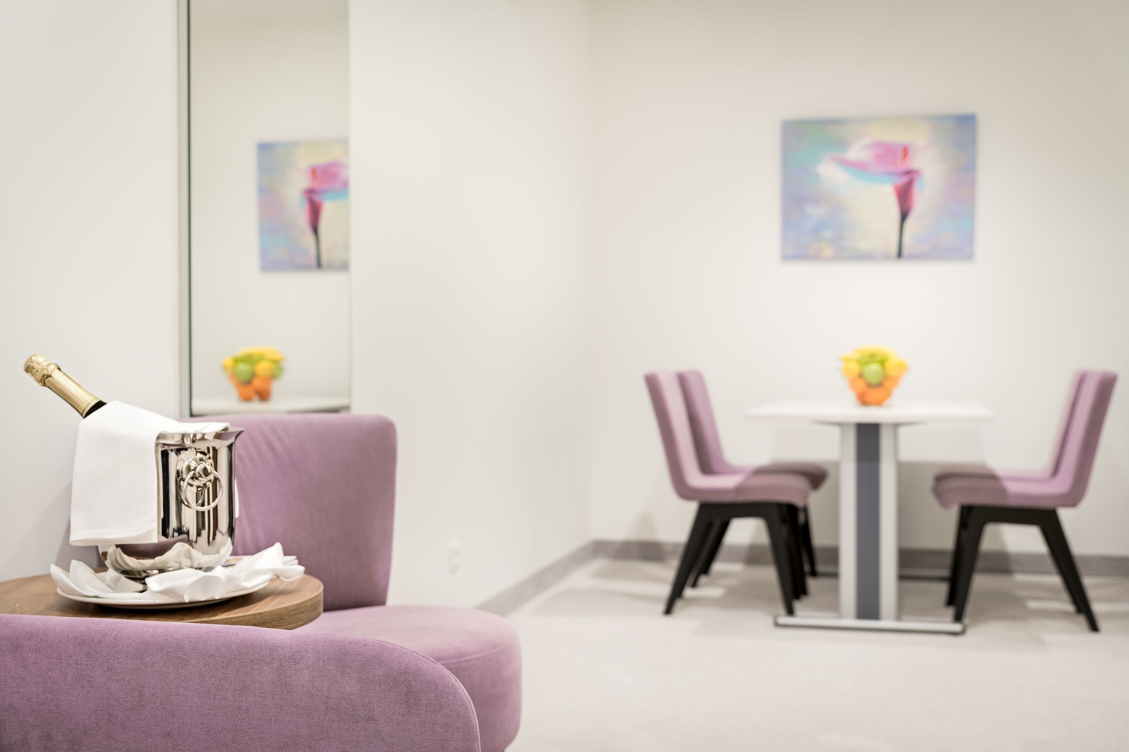 A modern room with a purple sofa, a small table with a bottle and glasses, and a dining area with a white table and purple chairs.