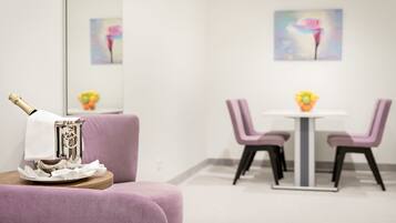 A modern room with a purple sofa, a small table with a bottle and glasses, and a dining area with a white table and purple chairs.