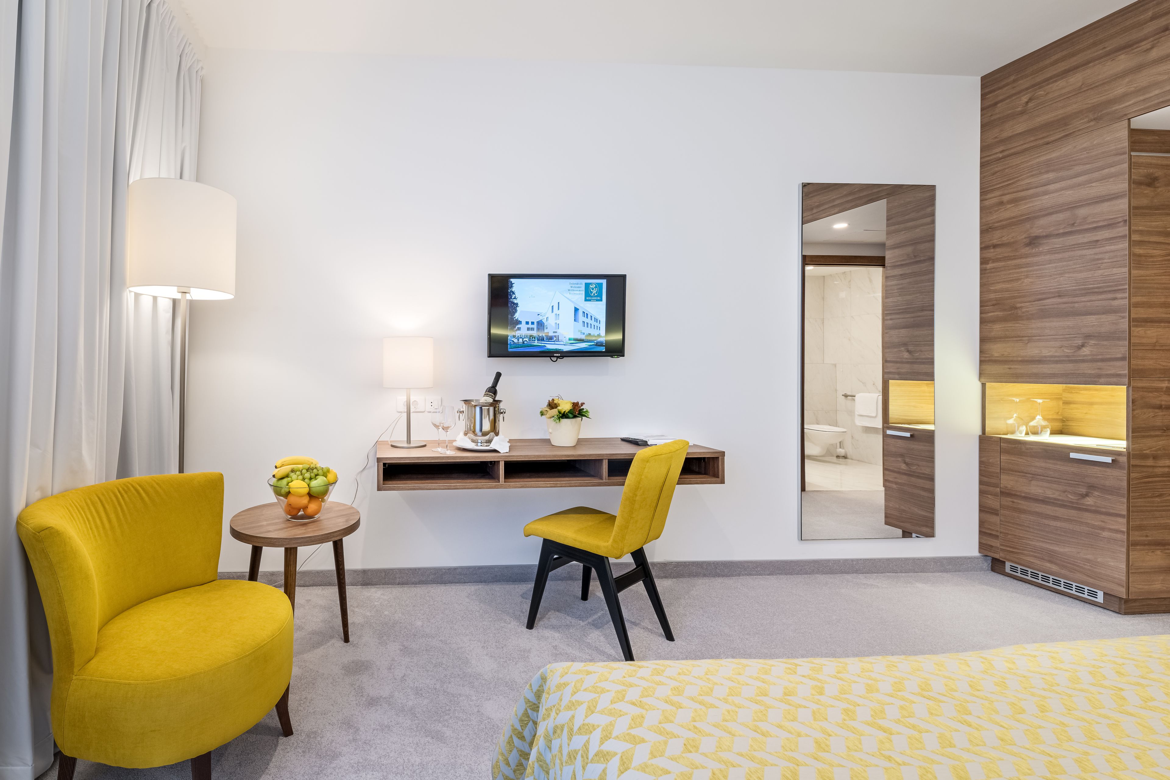 Habitación de hotel con cama, escritorio con computadora, silla, pequeña mesa con frutero, lámpara y televisión en la pared.