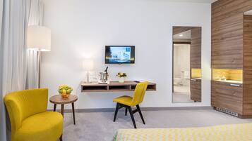 Habitación de hotel con cama, escritorio con computadora, silla, pequeña mesa con frutero, lámpara y televisión en la pared.