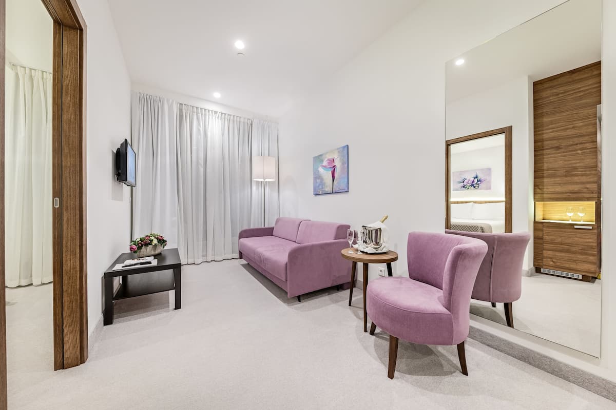 Ein modernes Hotelzimmer mit einem violetten Sofa, einem kleinen Tisch mit Blumen, einem an der Wand montierten Fernseher und einem Holzkabinett mit Spiegel.