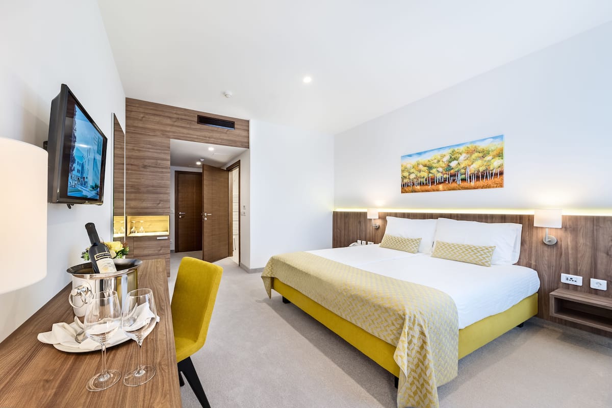 Ein Hotelzimmer mit einem großen Bett, einem an der Wand befestigten Fernseher, einem kleinen Tisch mit einer Champagnerflasche und Gläsern sowie einem Bild an der Wand.