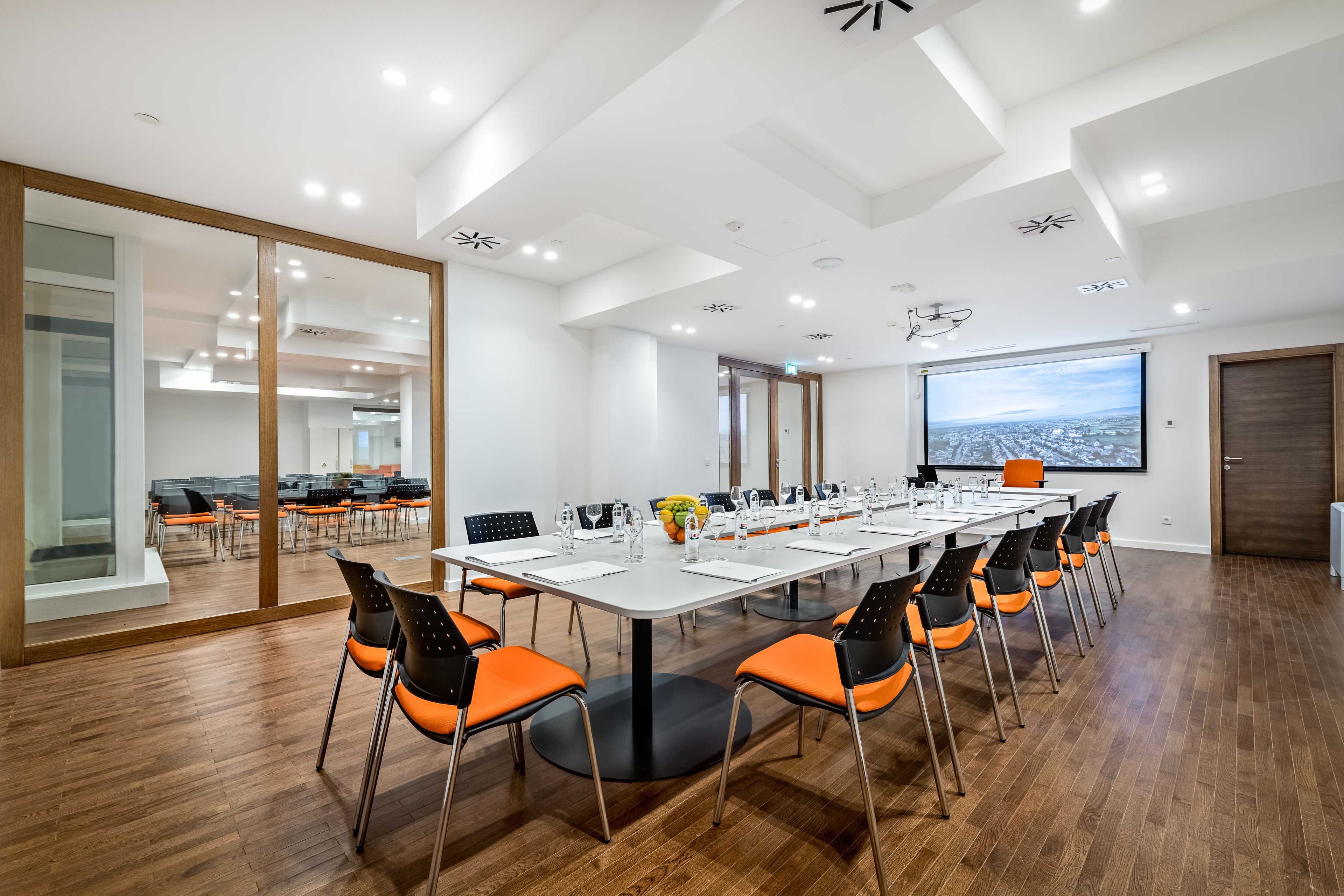 A modern conference room with a long table, orange chairs, and a large screen.