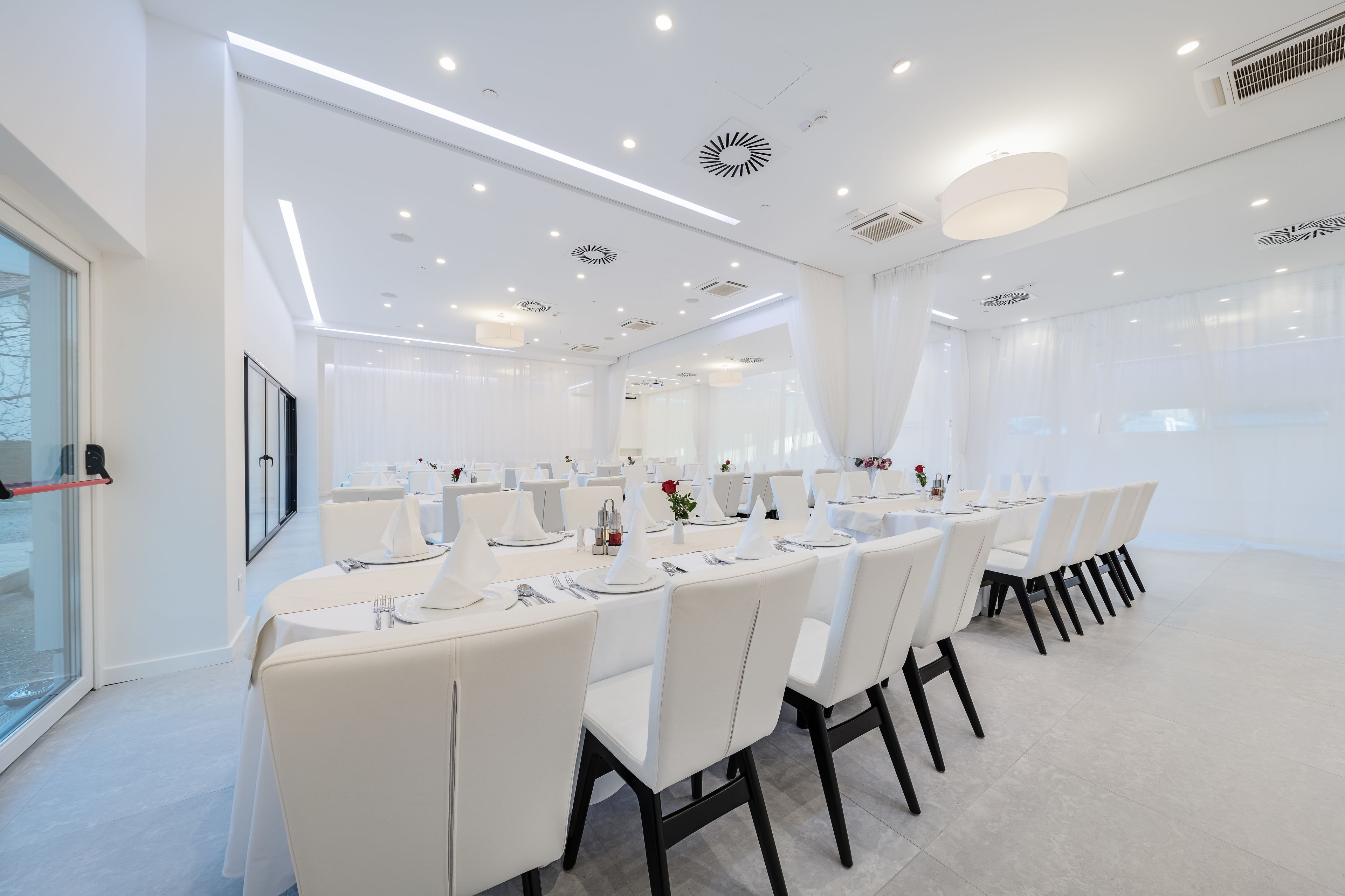 Een moderne eethoek met witte tafels en stoelen, inbouwspots en grote ramen met lichte gordijnen.