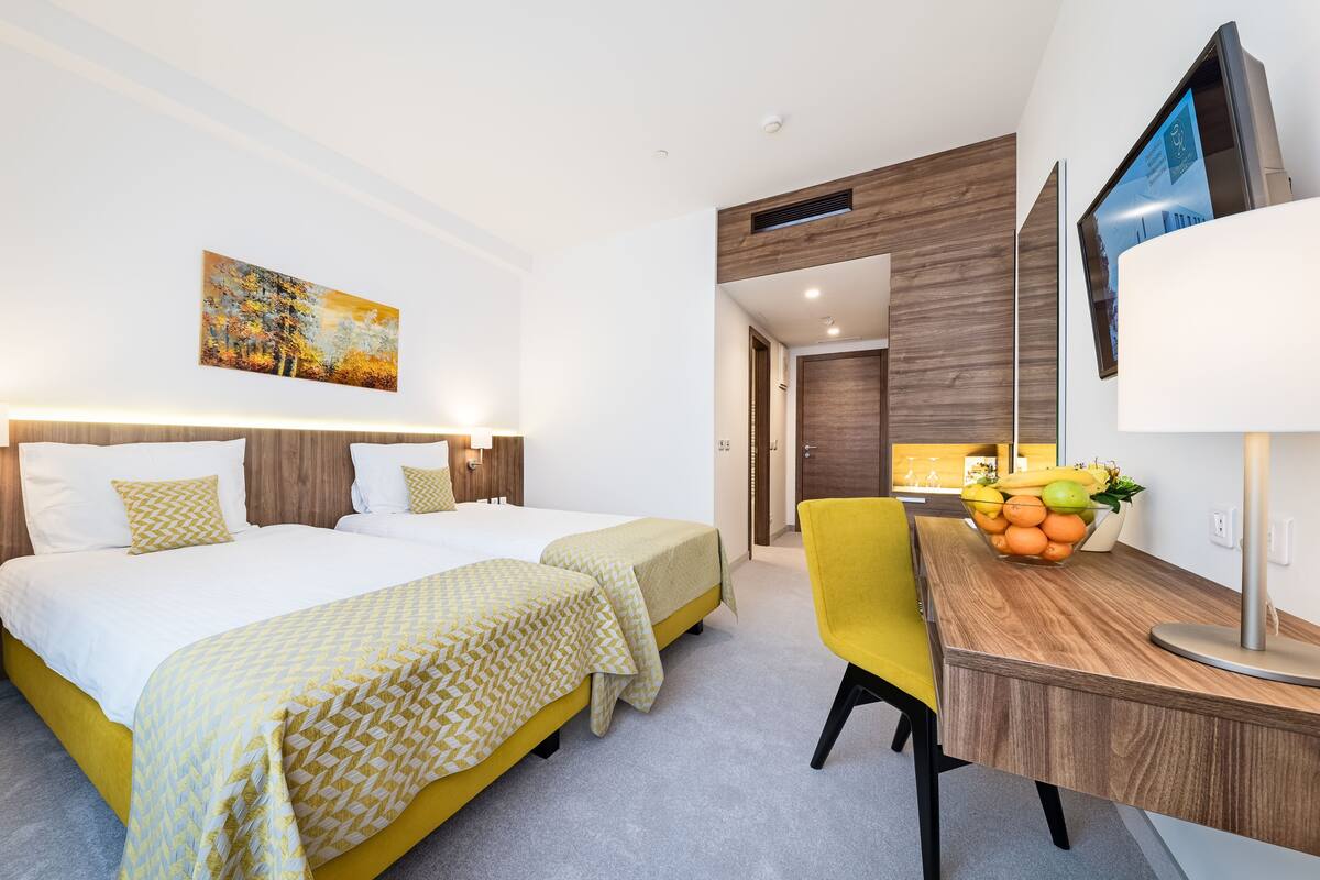 Ein Hotelzimmer mit zwei Betten, einem Schreibtisch mit Obstschale, einem Fernseher und einem Bild an der Wand.