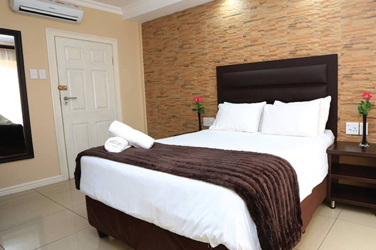 A hotel room with a large bed, a white door, a stone-patterned wall, and a nightstand with a vase of flowers.