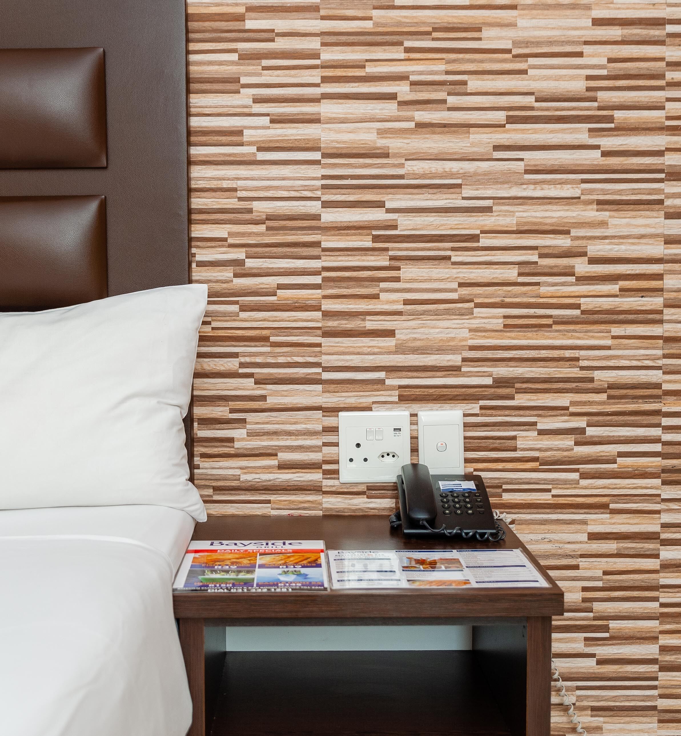 Hotelkamer met een wand met houtstructuur, een bed met wit bedlinnen, een nachtkastje met telefoon en brochures, en een stopcontact.