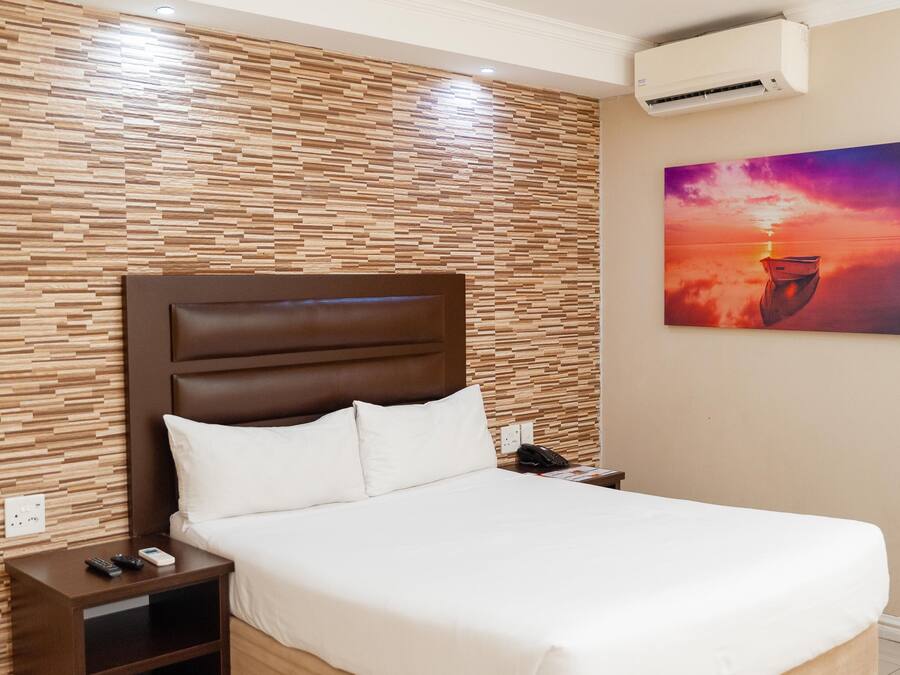 Quarto de hotel com parede de painel de madeira, cama com roupa branca, mesa de cabeceira com telefone e controles remotos e um aparelho de ar condicionado.