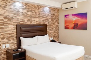 Quarto de hotel com parede de painel de madeira, cama com roupa branca, mesa de cabeceira com telefone e controles remotos e um aparelho de ar condicionado.