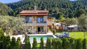 Una casa de dos plantas con piscina, rodeada de exuberante vegetación y una ladera boscosa.