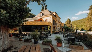 An outdoor café area with wooden benches and tables, surrounded by potted plants and a building with a balcony. - Hotel-Restaurant Le Cigalon (Waldbillig)