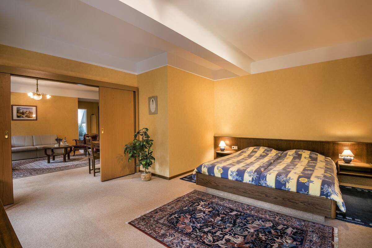 Ein Schlafzimmer mit einem großen Bett, Nachttischlampen, einem hölzernen Kopfteil, einem Beistelltisch, einer Topfpflanze und Blick auf das Wohnzimmer.