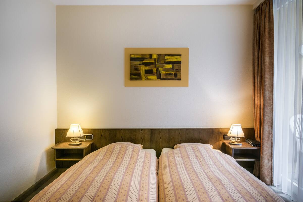 Ein Hotelzimmer mit zwei Betten, einem Kopfteil, einem Wandbild, Nachttischlampen und einem Fenster mit Vorhängen.