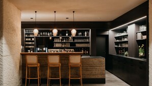 A bar area with three wooden bar stools, shelves with glasses and bottles, and pendant lighting. - Hotel-Restaurant Le Cigalon (Waldbillig)