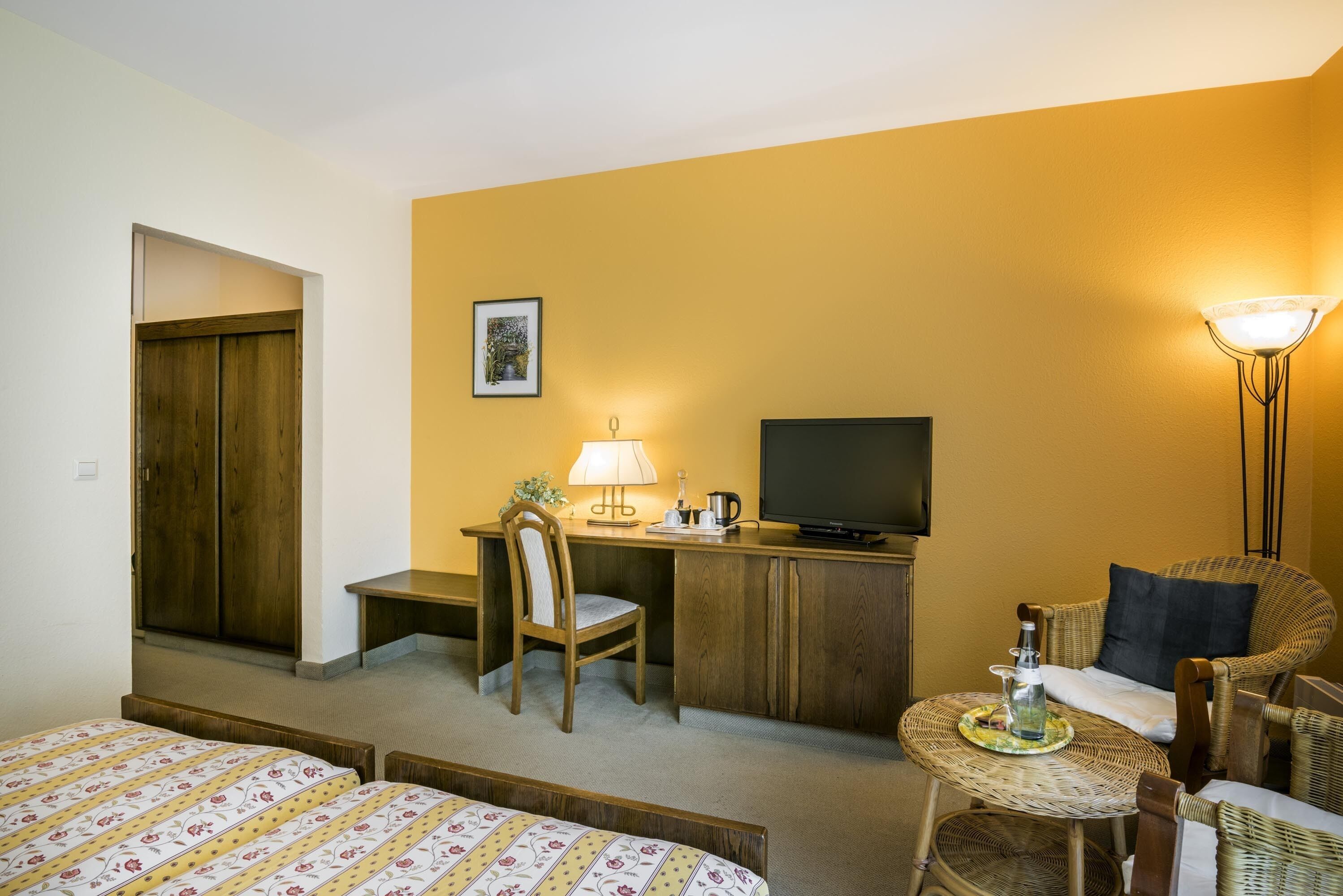 A hotel room with a bed, a desk with a chair, a television, a lamp, and a small table with a bottle and glasses.