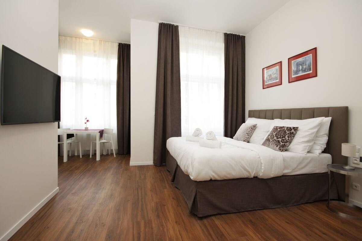 A hotel room with a bed, a TV, a desk, and two framed pictures on the wall.