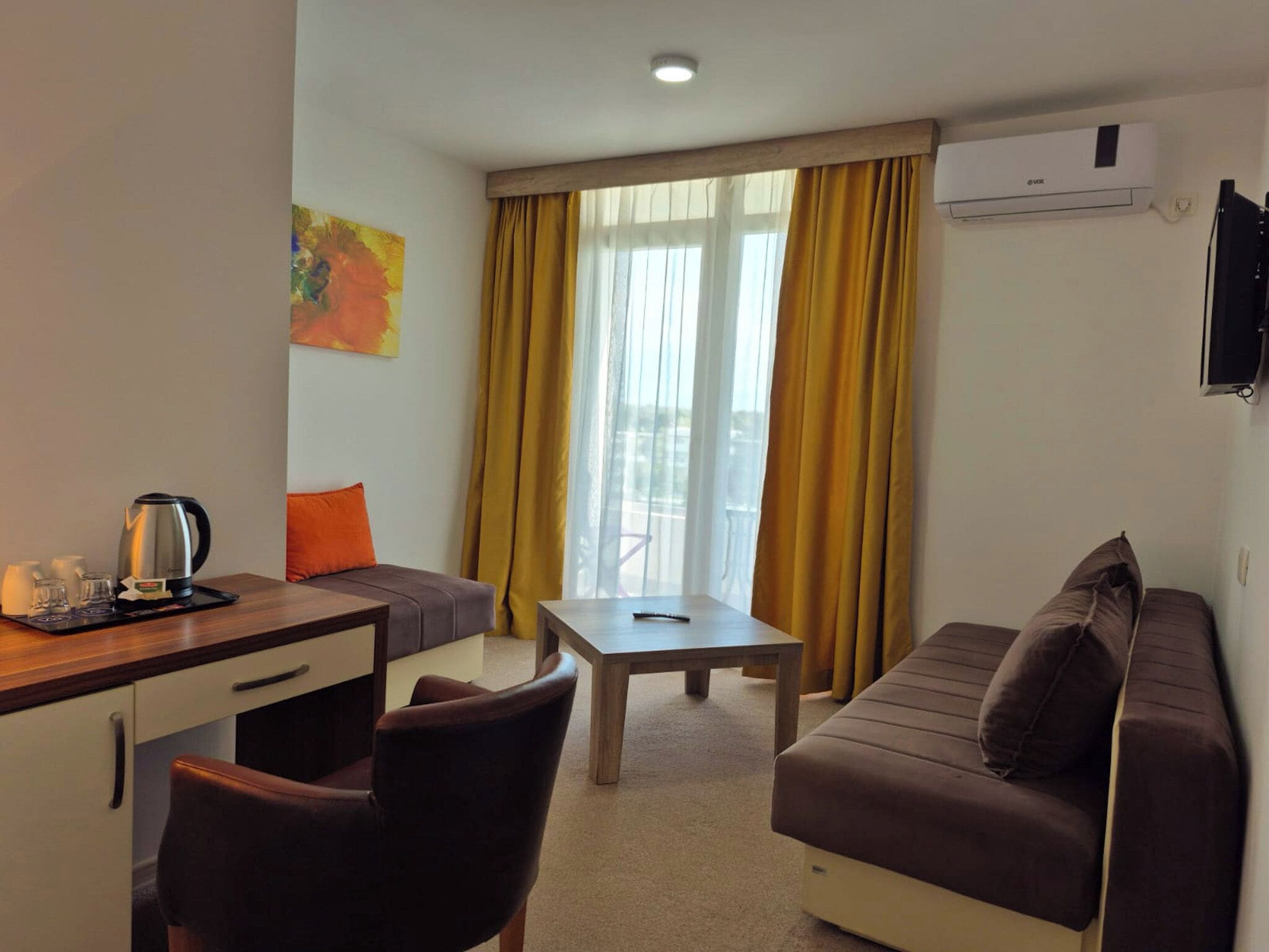 Quarto de hotel compacto com sofá, pequena mesa, escrivaninha e cadeira. Há uma janela com cortinas amarelas, um aparelho de ar condicionado na parede e um quadro na parede.
