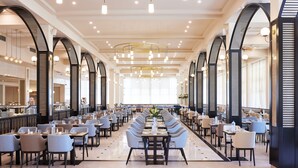 Un vaste espace dédié à la salle à manger, avec de nombreuses tables et chaises, de hauts plafonds dotés de luminaires modernes et de grandes fenêtres laissant entrer la lumière naturelle.