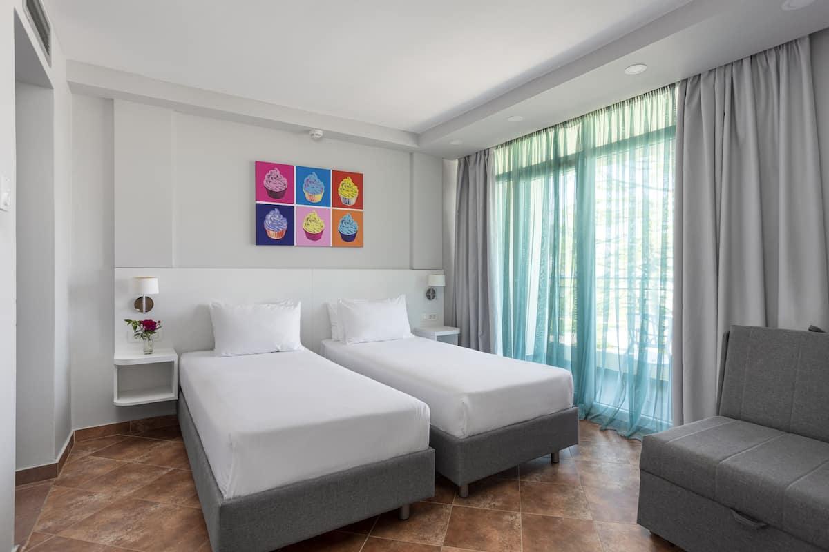 Een hotelkamer met twee enkele bedden, een grijze bank, een wanddecoratie van cupcakes, een raam met dunne gordijnen en een klein nachtkastje met een lamp en vaas.