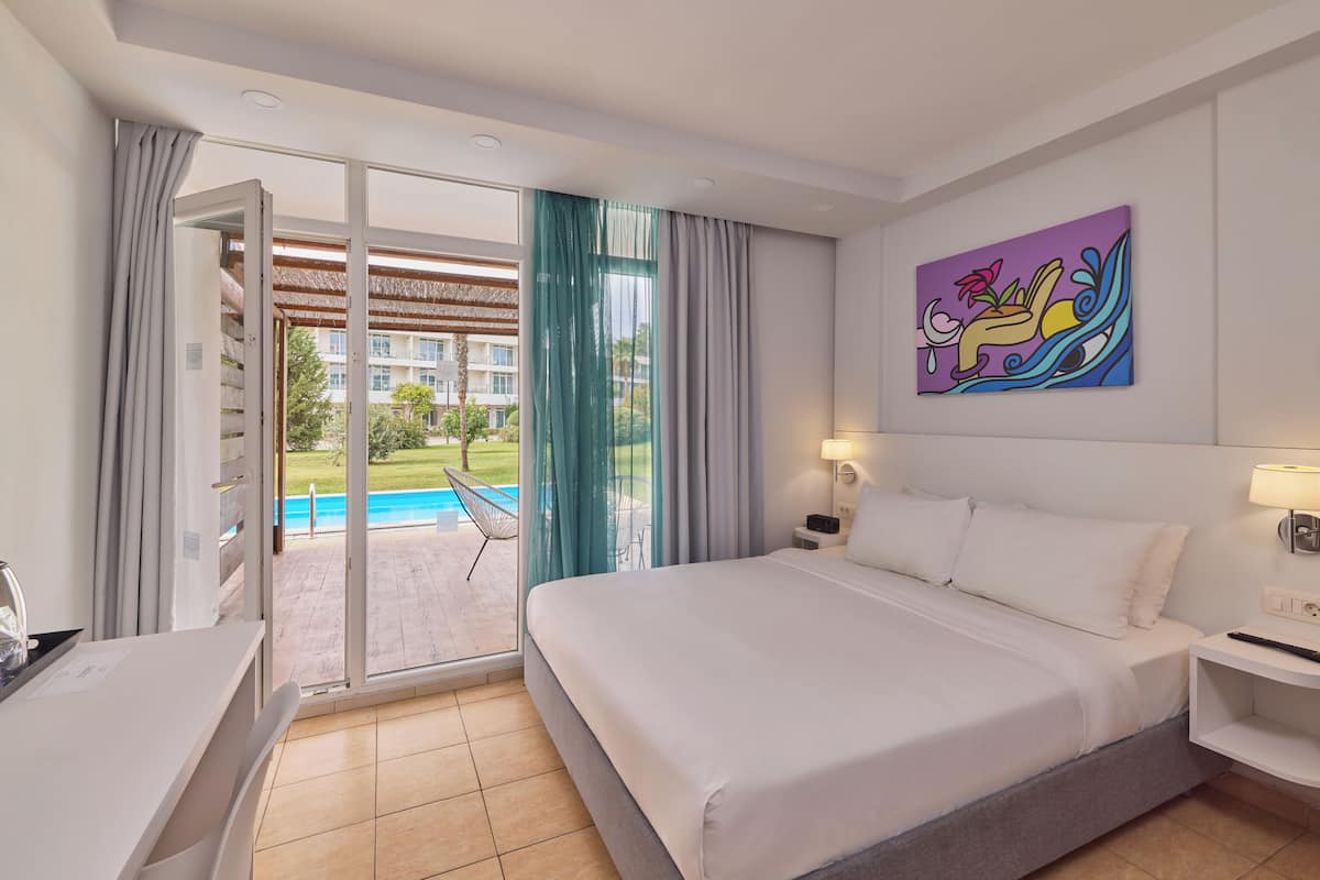 Een hotelkamer met een bed, bureau, stoel, schilderij en uitzicht op een zwembad en groen.