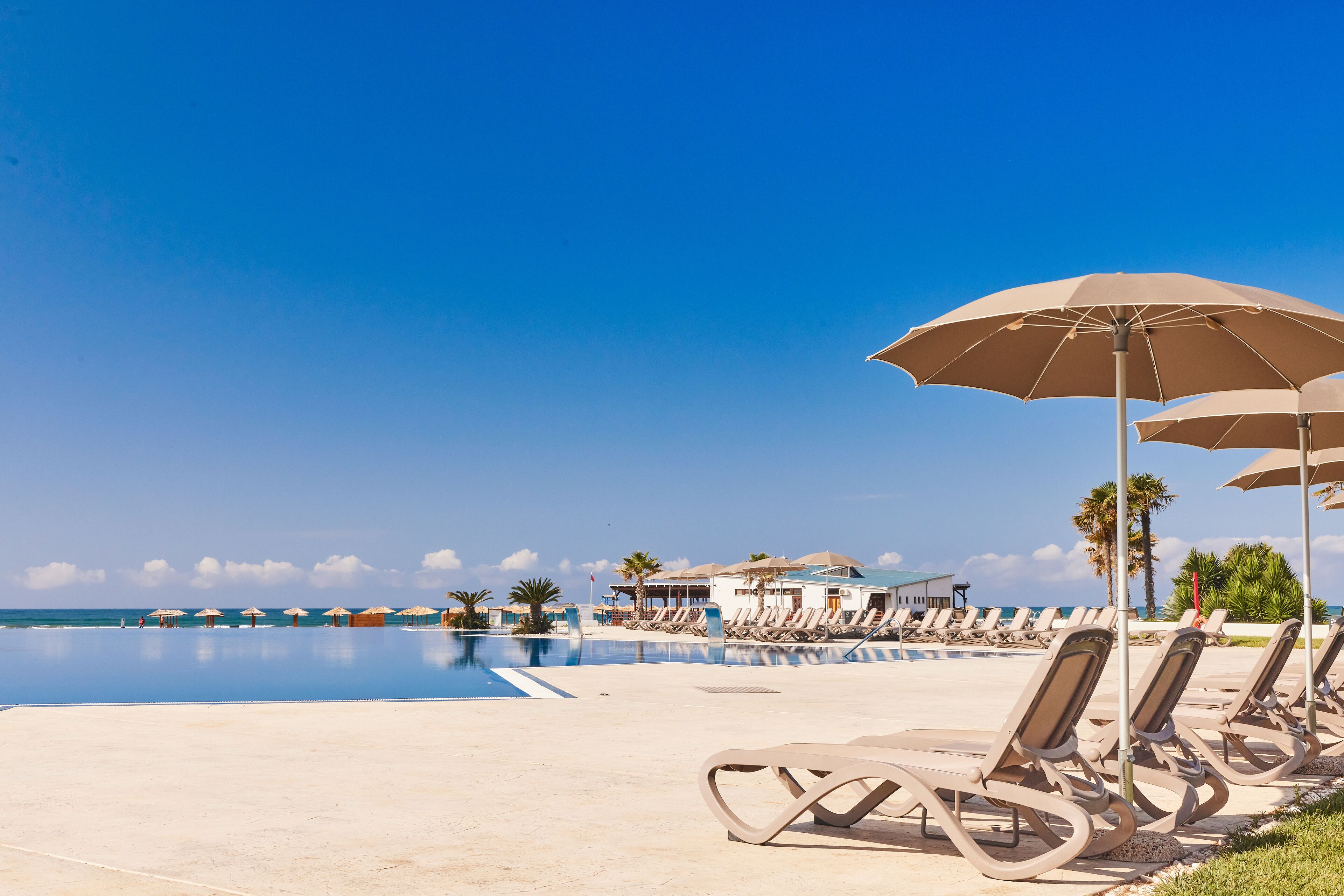 A poolside area with lounge chairs and umbrellas, a clear blue sky, and a building in the background.