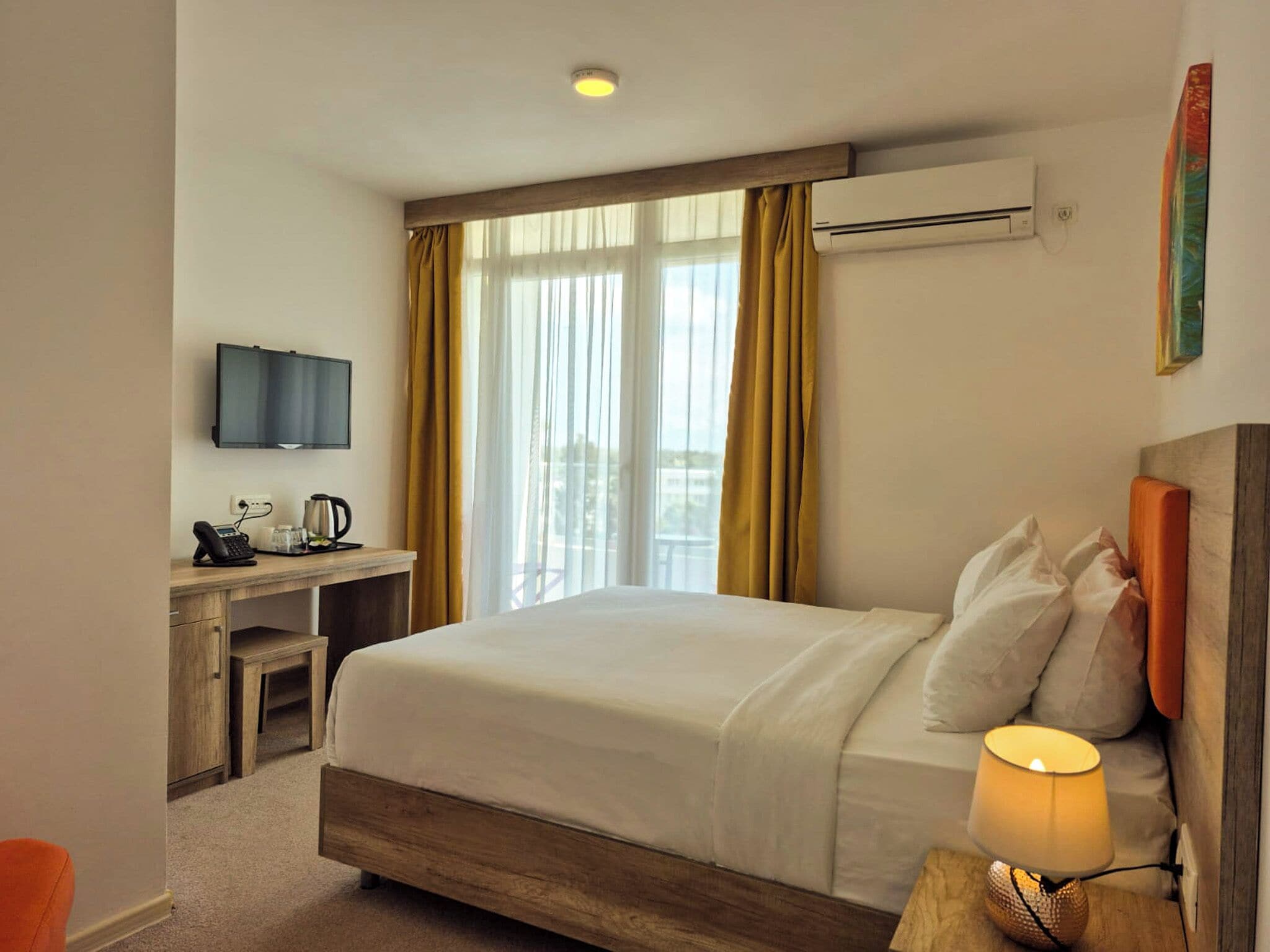 Habitación de hotel con cama, escritorio con teléfono y cafetera, televisión montada en la pared y ventana con cortinas.